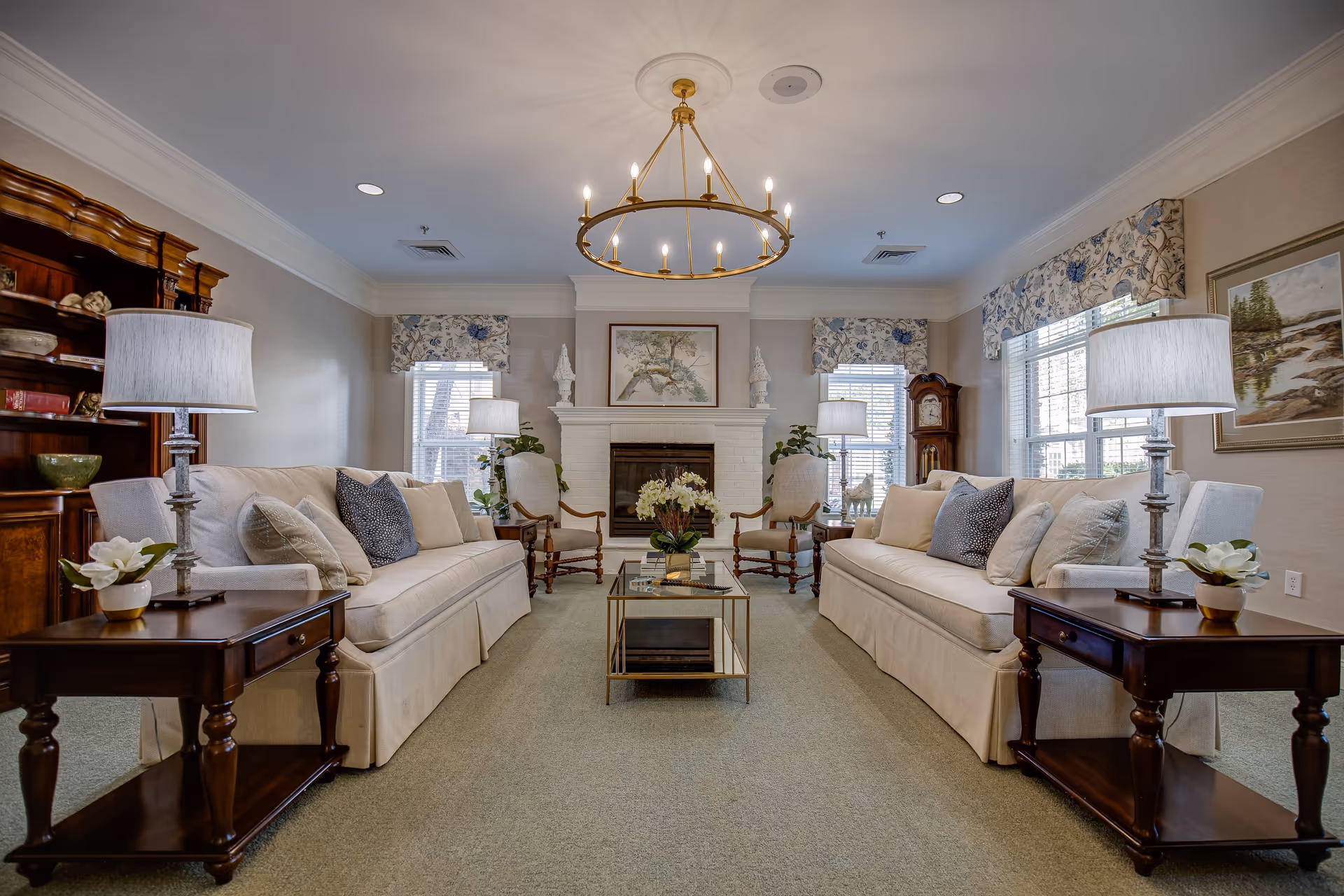 A cozy living room with two beige sofas facing each other, each adorned with multiple cushions. Between the sofas is a glass coffee table with a flower arrangement. On either side of the sofas are wooden side tables with lamps and small flower pots. The room features a white brick fireplace with decorative sculptures and a framed painting above it. Two armchairs are placed near the fireplace, and large windows with floral valances allow natural light to fill the room. A wooden grandfather clock and a wooden bookshelf are also visible.