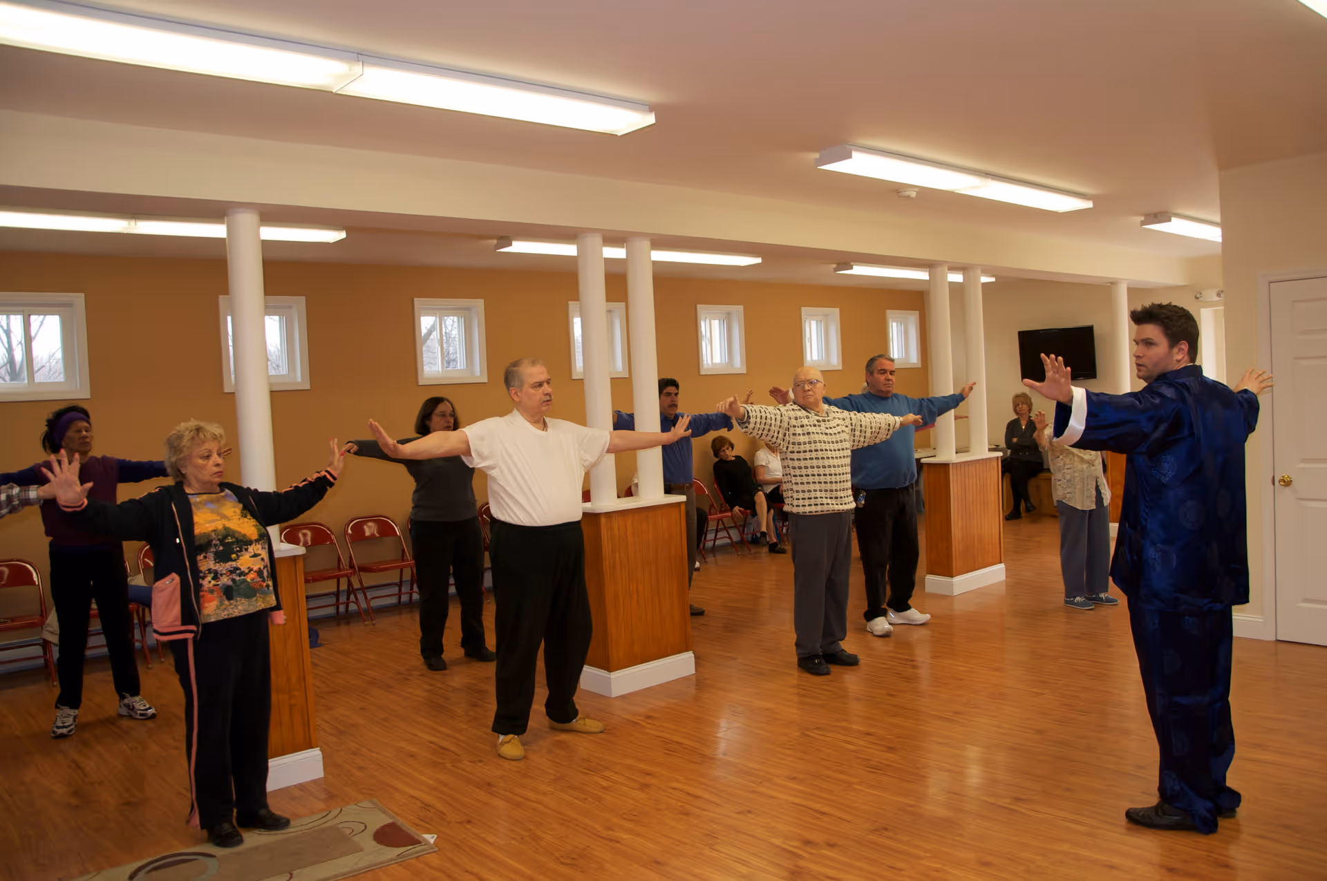 A group of elderly people standing in a spacious room with wooden floors and beige walls, participating in a guided exercise or stretching session led by an instructor wearing a blue outfit. The participants have their arms extended to the sides.