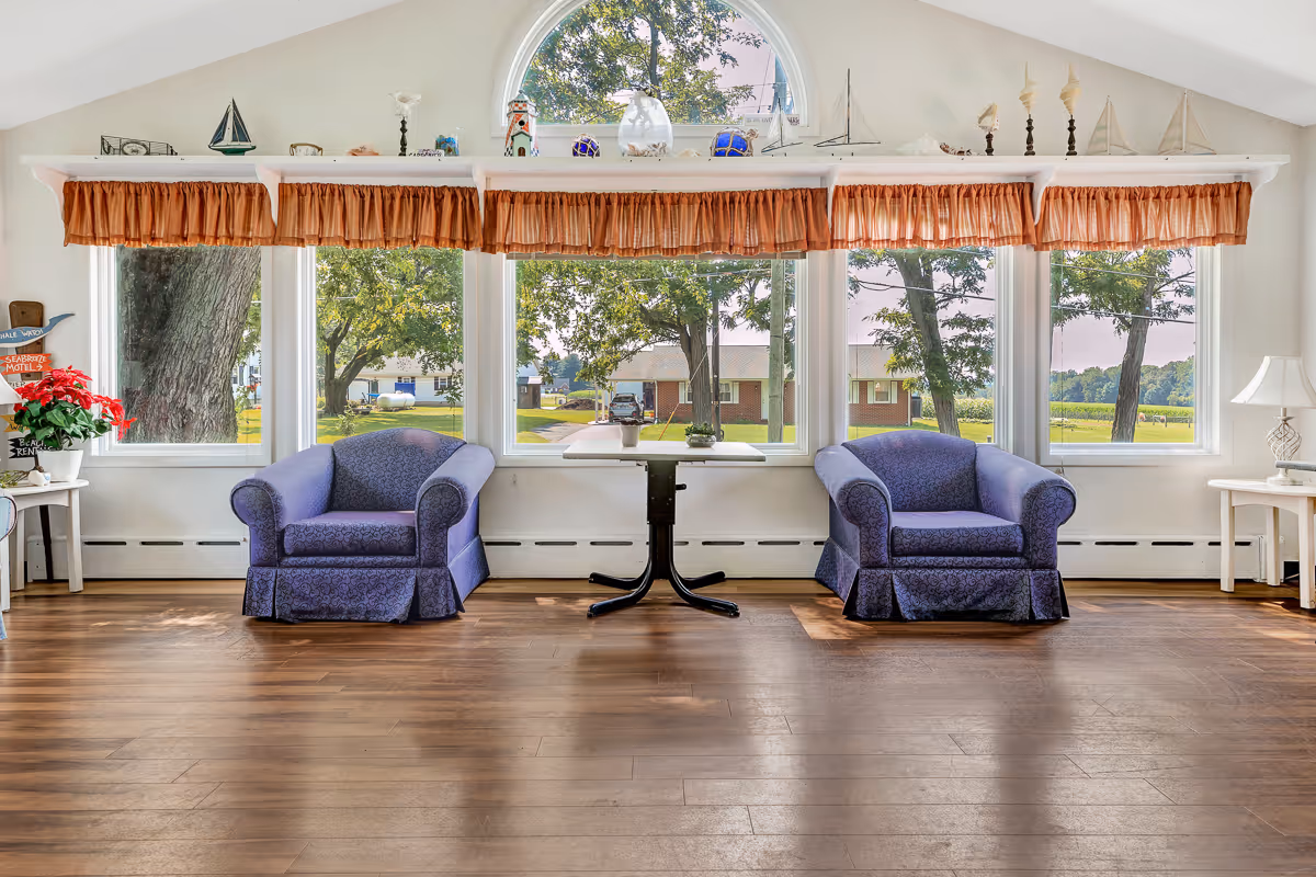 Sunlit sitting area with two purple armchairs and a small table in front of large windows looking out to trees and a lawn.
