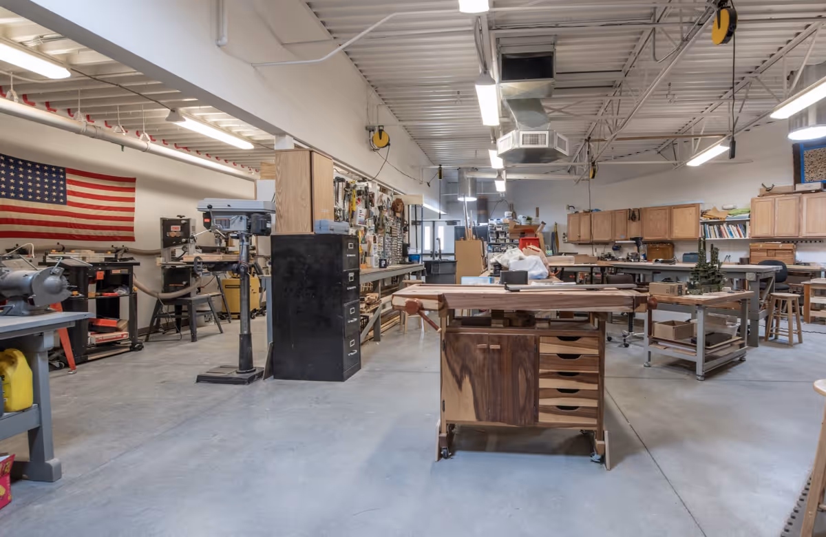 A spacious workshop area with various woodworking tools and equipment, including drill presses, workbenches, and storage cabinets. An American flag is displayed on the wall, and the room has a high ceiling with exposed ductwork and fluorescent lighting.