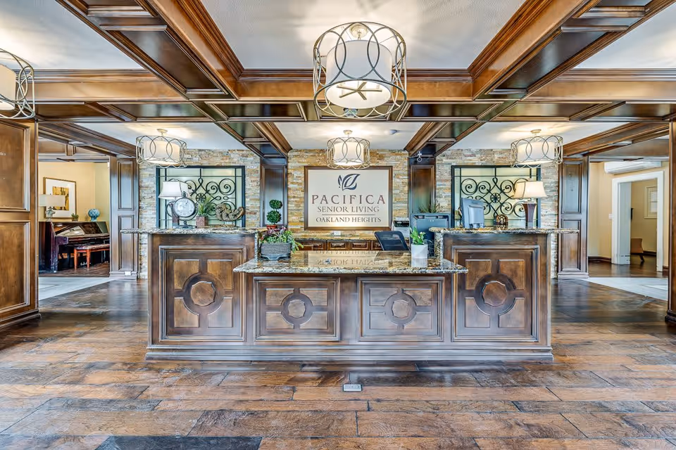 Reception area of a senior living facility with a large wooden front desk featuring granite countertops. Behind the desk is a sign that reads 'Pacifica Senior Living Oakland Heights'. The room has wooden coffered ceilings, hanging light fixtures, and decorative plants on the desk. There are open doorways on either side leading to other rooms.
