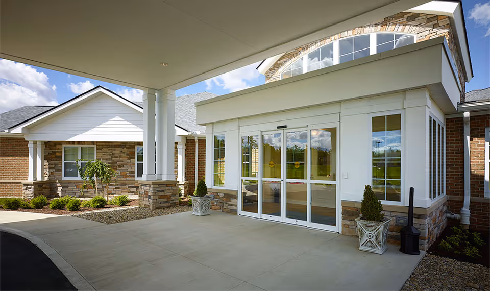 Covered porte-cochère and glass entrance to a brick-and-stone senior living building with planters and landscaping.