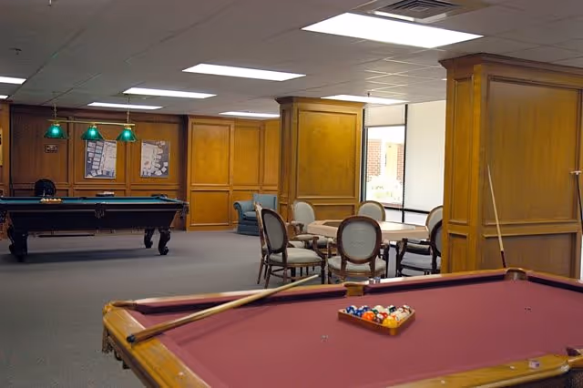 Interior view of a recreational room with two pool tables, one in the foreground with a cue stick and balls arranged, and another in the background under green hanging lights. There is a round table with chairs and wood-paneled walls with windows letting in natural light.