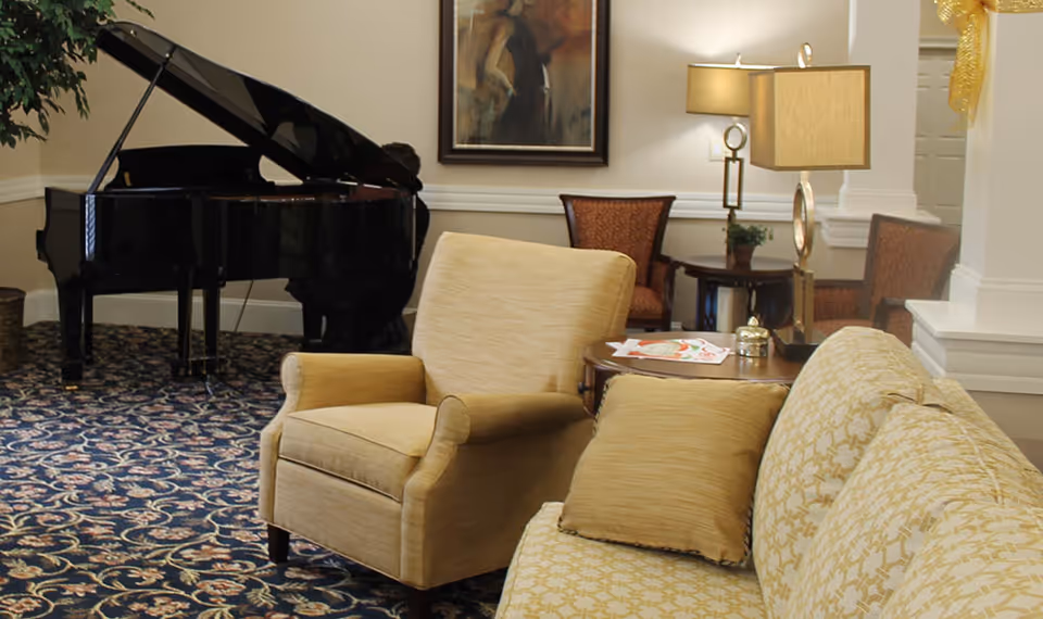 A cozy living room area with a beige armchair and matching sofa with patterned cushions. A round wooden side table with a lamp and some papers is placed between the armchair and sofa. In the background, there is a black grand piano, two upholstered chairs, a small round table with a lamp, and a framed painting on the wall. The room has patterned carpet flooring and white walls with molding.