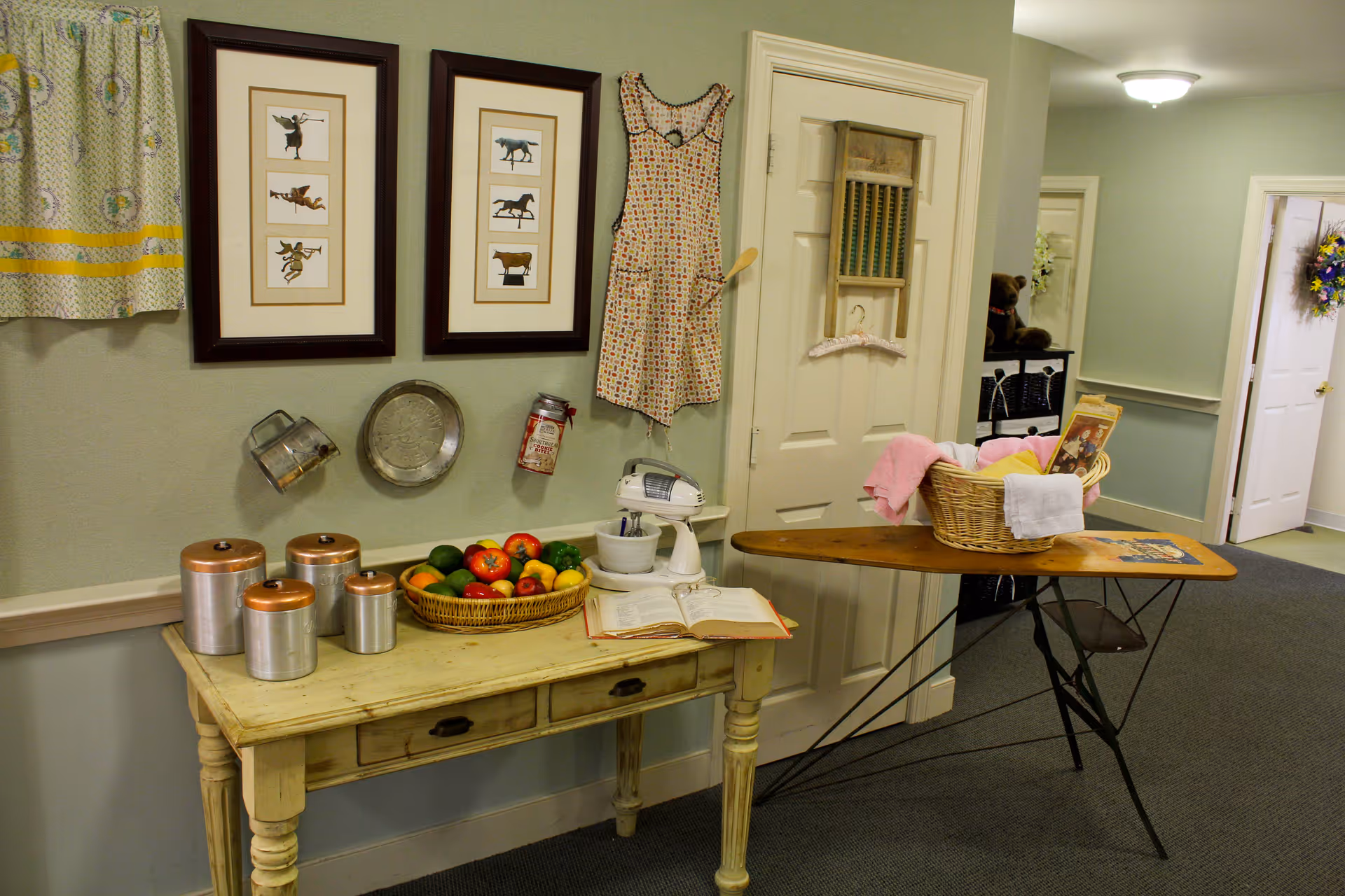 A cozy interior corner of a senior living facility featuring a vintage-style wooden table with copper canisters, a basket of artificial fruits, an open book, and a white mixer. Above the table are two framed pictures of animals, a hanging apron, and some kitchen utensils on the wall. To the right, there is an ironing board with a basket containing towels and a magazine. The hallway in the background has doors and a floral wreath on one door.