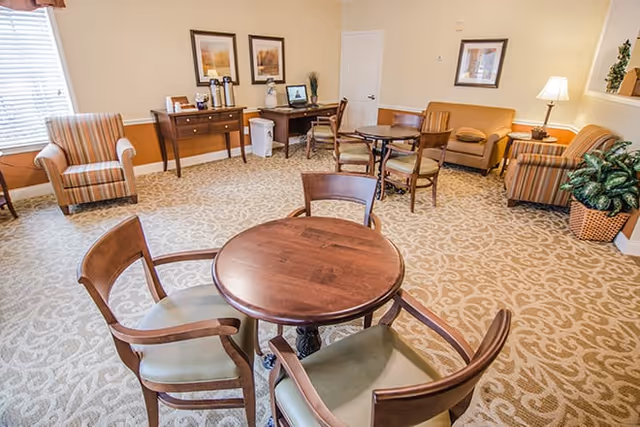 A cozy common area with patterned carpet, featuring a round wooden table with four chairs in the foreground. In the background, there are two armchairs, a small sofa, a side table with a lamp, a console table with coffee dispensers, and framed artwork on the walls. A potted plant is placed near the corner.