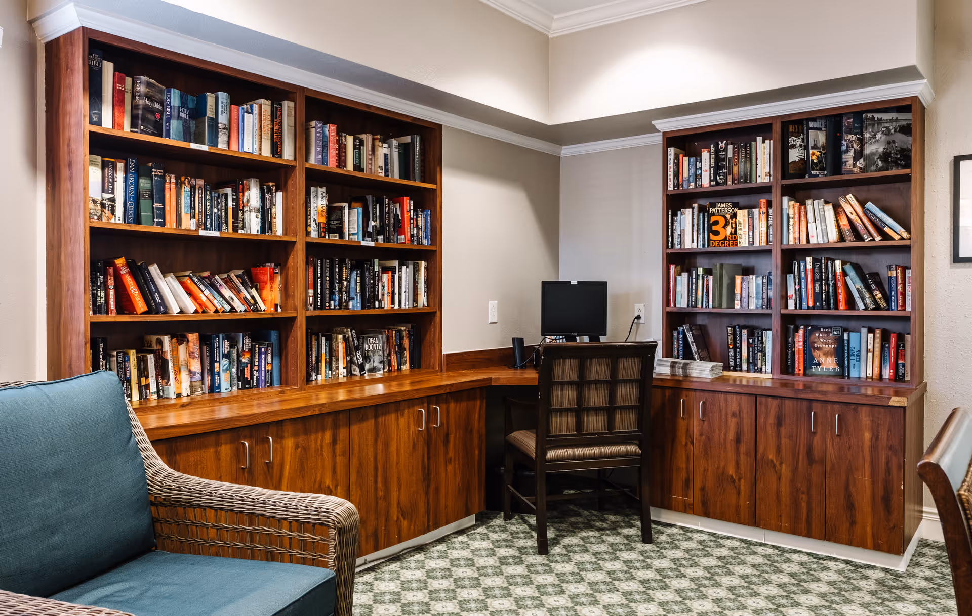 A cozy reading and computer area with wooden bookshelves filled with books, a computer on a wooden desk, a chair in front of the desk, and a wicker armchair with a blue cushion. The room has a patterned carpet and neutral-colored walls.