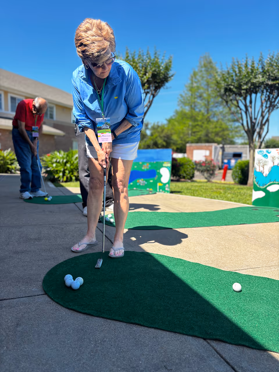 An elderly woman wearing a blue jacket and white shorts is playing mini golf outdoors on a sunny day. She is holding a putter and aiming at a golf ball on a green artificial turf. In the background, an elderly man in a red shirt is also playing mini golf. Trees, bushes, and a building are visible under a clear blue sky.