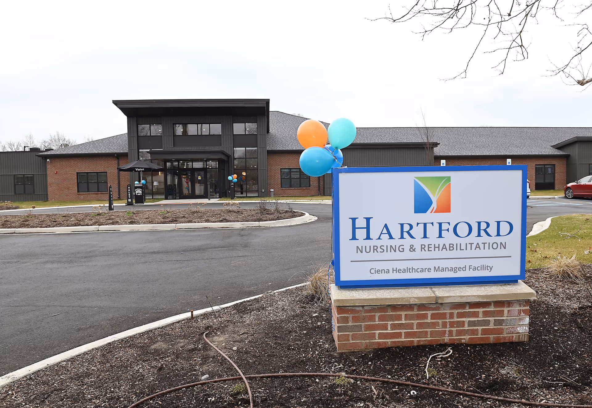 Exterior view of Hartford Nursing and Rehabilitation facility with a large sign in the foreground displaying the facility's name and logo. The building is modern with brick and dark paneling, and there are colorful balloons attached near the entrance.