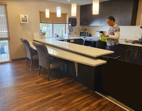 A senior woman standing in a modern kitchen area with dark cabinets and a white countertop. The kitchen features a microwave, coffee maker, and several cups on the counter. There are three chairs along the counter and pendant lights hanging from the ceiling. A window with blinds is visible in the background.