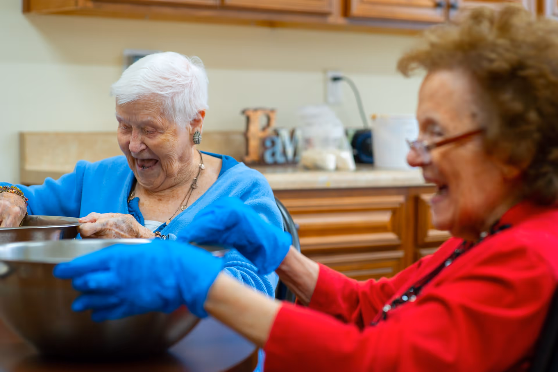 Two elderly women sitting at a table in a kitchen, smiling and engaging in an activity with large metal mixing bowls. One woman is wearing a blue sweater and the other a red sweater with blue gloves.