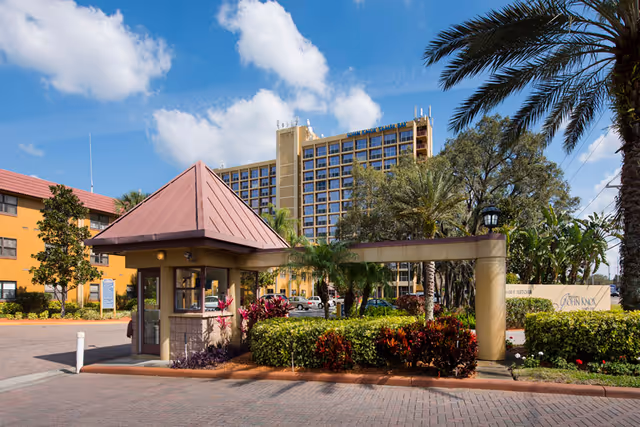Entrance gatehouse with a small guard booth featuring a red sloped roof, surrounded by landscaped bushes and palm trees. Behind the gatehouse, there is a tall multi-story building and a shorter yellow building under a blue sky with some clouds. A sign near the bushes reads 'John Knox Village'.