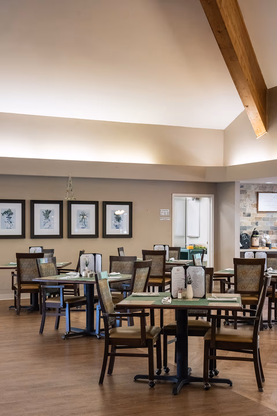 A dining room with several tables and chairs arranged neatly. Each table has placemats, menus, and small decorative plants. The walls are adorned with framed botanical prints, and the ceiling features wooden beams. The room has a warm and inviting atmosphere with soft lighting.