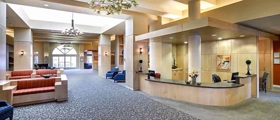 Spacious and well-lit reception area of Eskaton Village Carmichael featuring a large curved front desk with computer monitors, decorative plants, and flowers. The room has patterned carpet flooring, beige walls, and multiple seating areas with red and blue upholstered chairs and sofas. Large windows and a chandelier provide natural and artificial lighting.