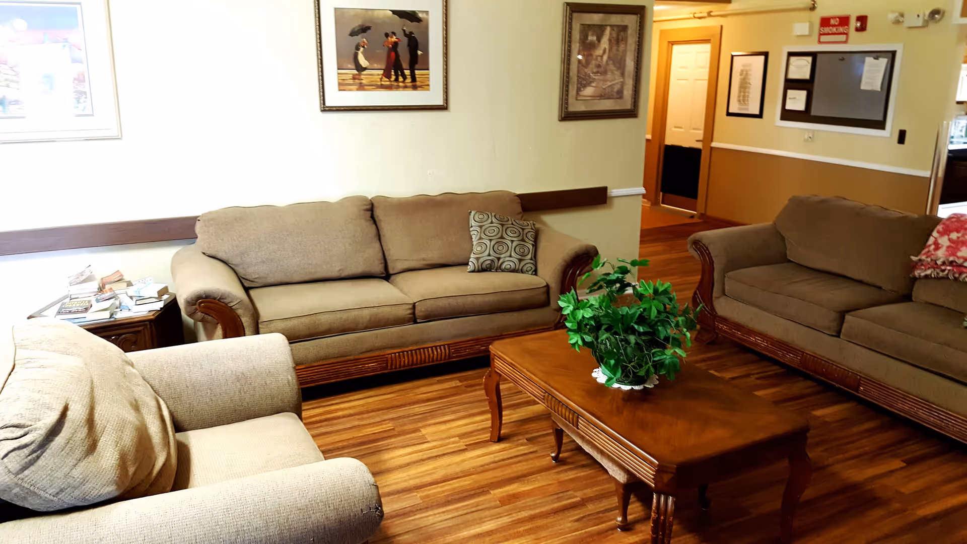 A cozy living room with two brown sofas and one beige armchair arranged around a wooden coffee table with a green potted plant. The room has wooden flooring, framed artwork on the walls, and a small side table with books stacked on it.