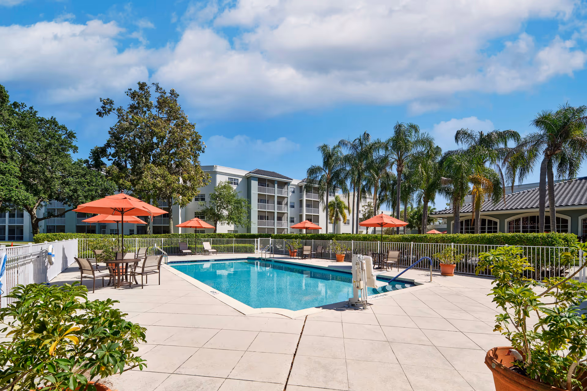 Outdoor swimming pool area with clear blue water surrounded by a tiled deck. Several tables with orange umbrellas and chairs are placed around the pool. Palm trees and other greenery are visible in the background along with a multi-story residential building and a clubhouse under a partly cloudy blue sky.