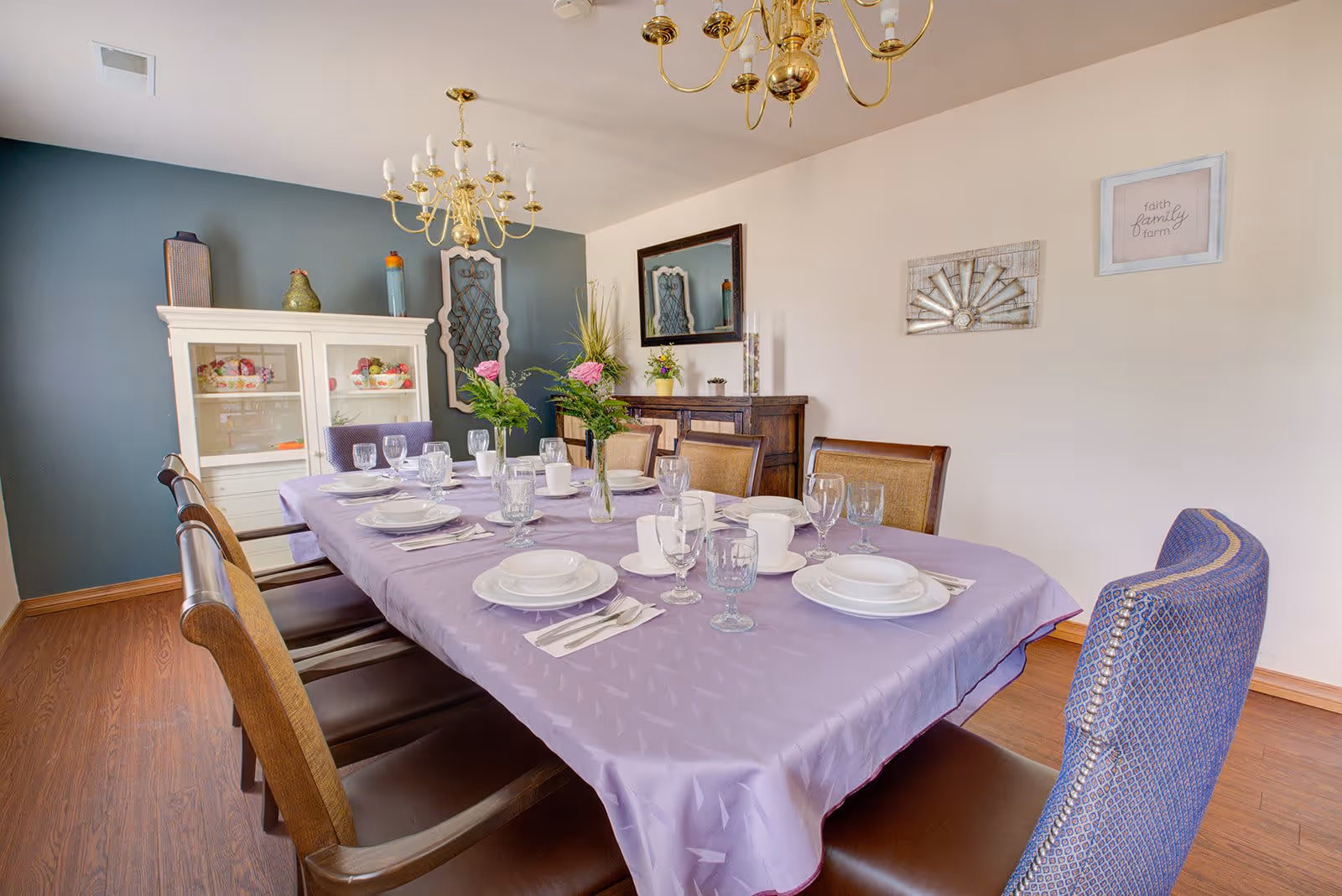 A dining room with a long table covered with a lavender tablecloth set with white plates, cups, and glassware. There are eight chairs around the table, a white cabinet with decorative items against a blue accent wall, a wooden sideboard with a mirror above it, and two chandeliers hanging from the ceiling. The walls have framed decorative art pieces.