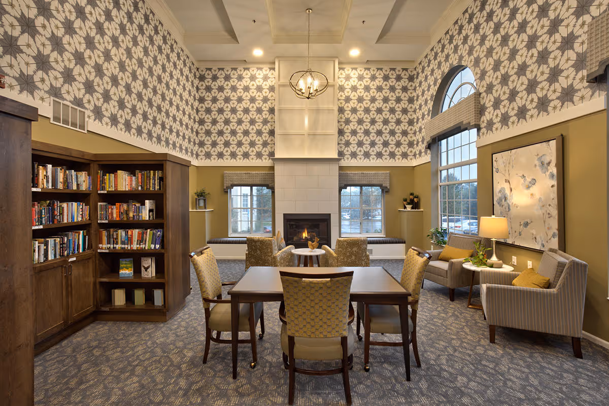 Communal living room and library with a central table and chairs, bookshelves, seating areas, a fireplace, and large windows under high patterned wallpaper.