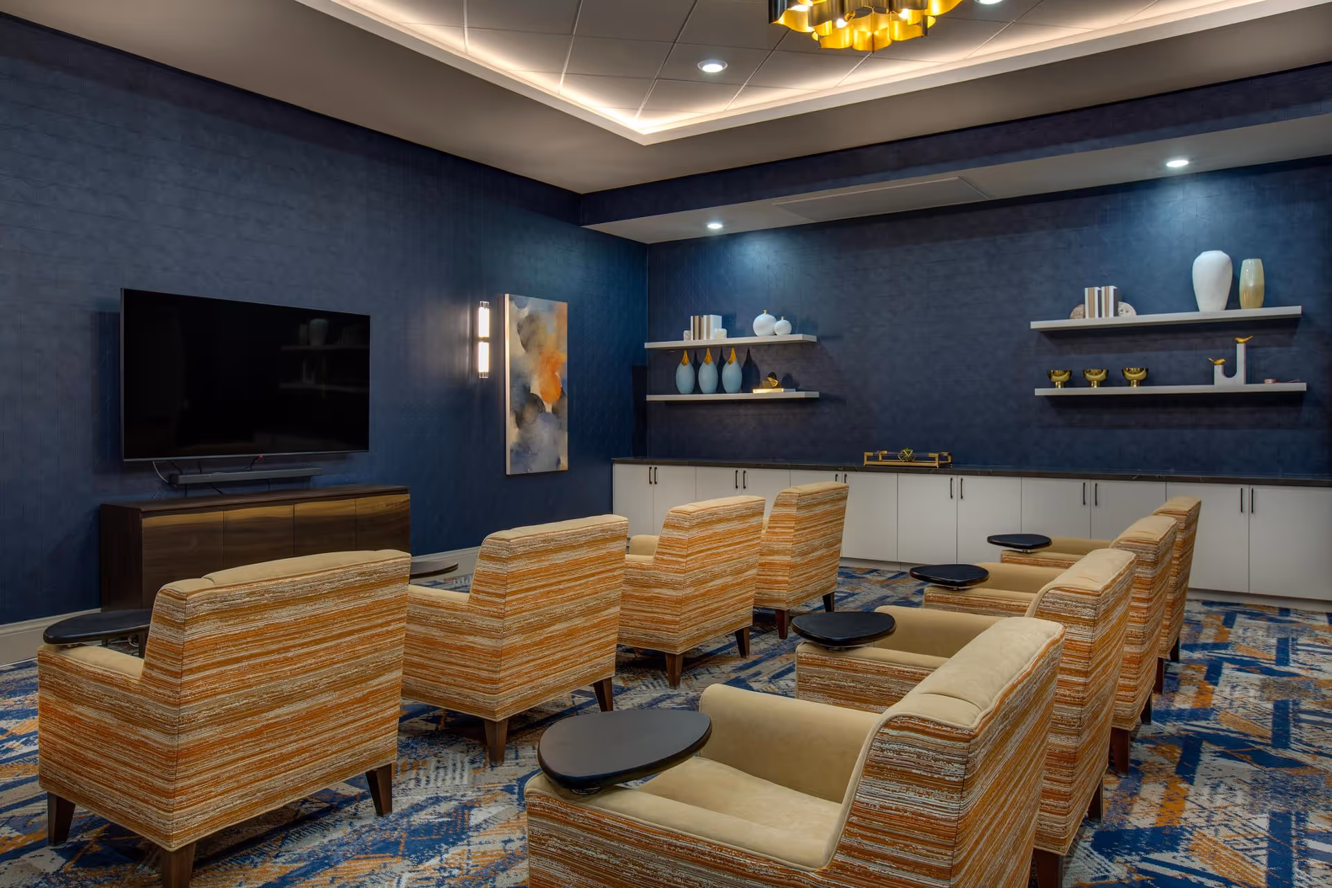A cozy media room with multiple striped armchairs arranged in rows facing a large flat-screen TV mounted on a dark blue wall. The room features a patterned carpet with blue and orange tones, white cabinets along the back wall, and decorative shelves with vases and small sculptures. The ceiling has recessed lighting and a modern chandelier.