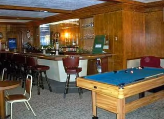 Interior view of a recreational room featuring a pool table with billiard balls, a bar area with several bar stools, wood-paneled walls, and carpeted floor.