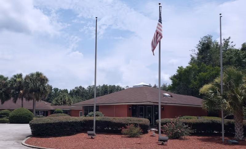 Single-story senior living building entrance with an American flag, trimmed hedges, palm trees, and a circular driveway.