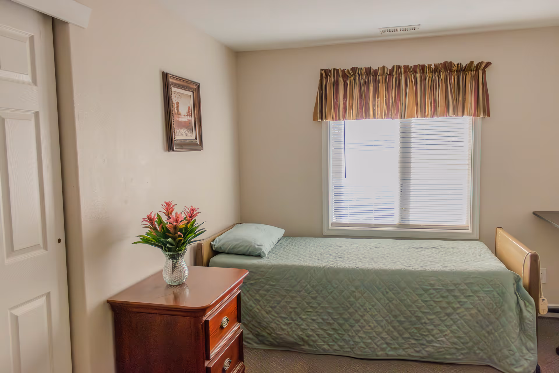 A small bedroom with a single bed covered in a green quilt and a matching pillow. Next to the bed is a wooden nightstand with two drawers and a glass vase holding pink flowers. A framed picture hangs on the beige wall above the nightstand. A window with white blinds and a striped valance lets in natural light.