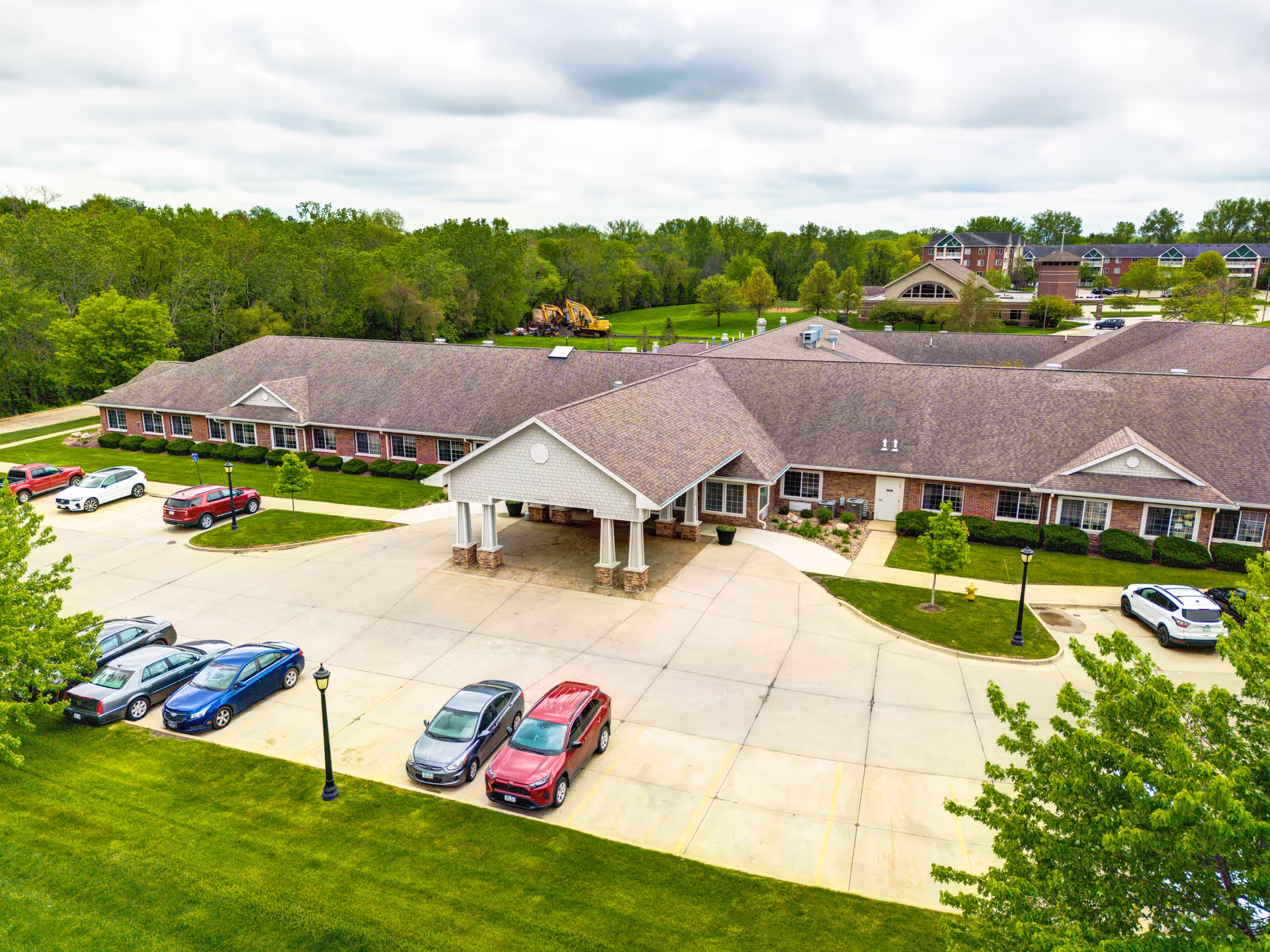 Aerial view of a single-story senior living facility building with a covered entrance and several parked cars in front. The building is surrounded by green lawns, trees, and a wooded area in the background under a cloudy sky.