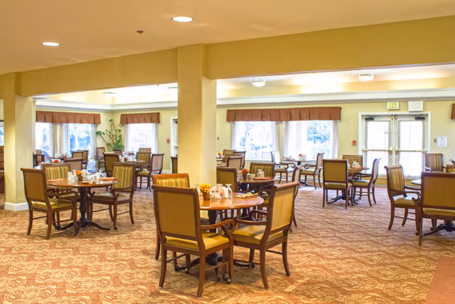 A spacious dining room with multiple round wooden tables and cushioned chairs arranged neatly. The room has large windows with brown valances allowing natural light to brighten the space. The carpet has a patterned design, and there are small floral centerpieces on each table. The walls are painted a light yellow color, and there are ceiling lights providing additional illumination.