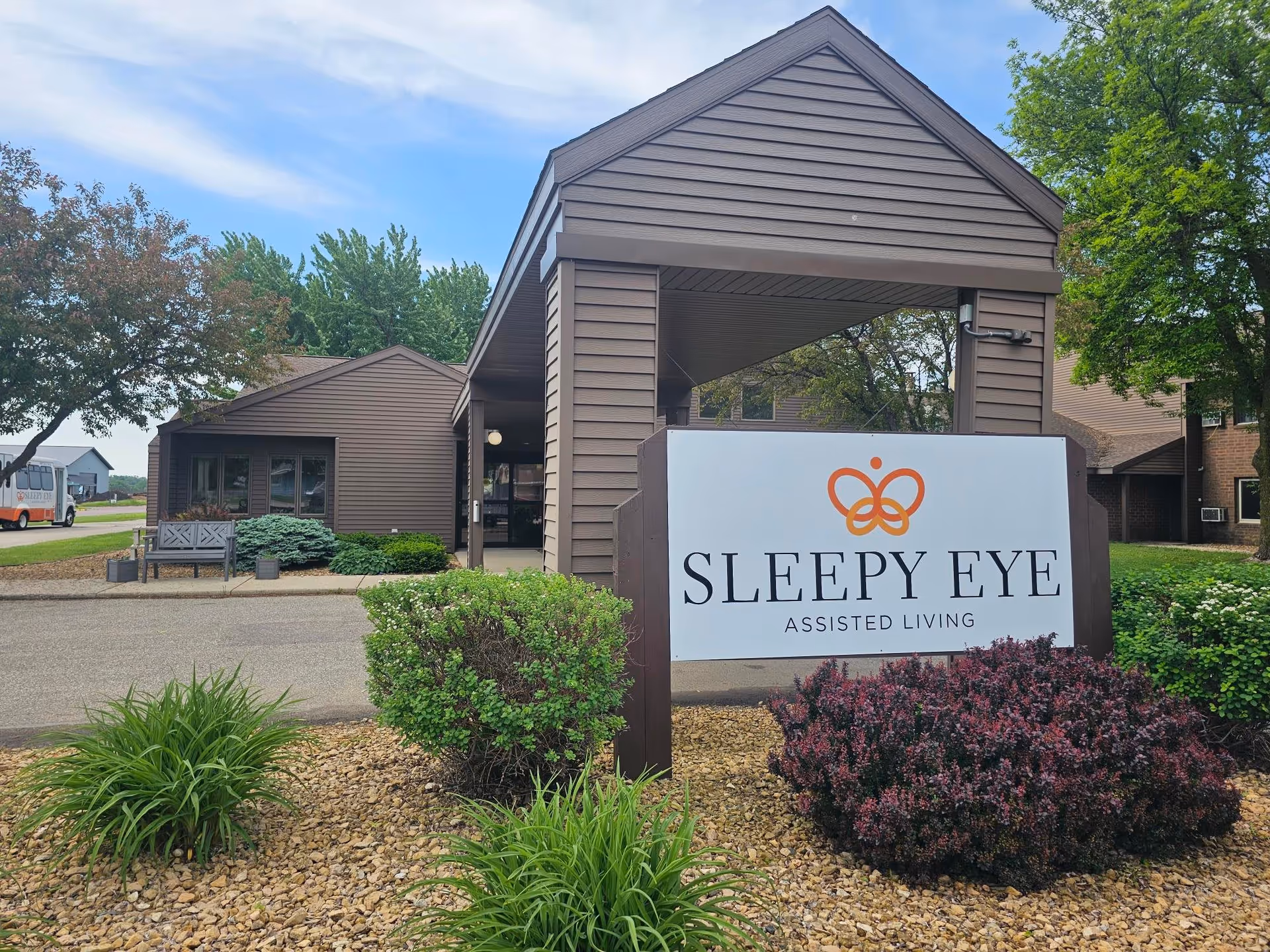 Exterior view of Sleepy Eye Assisted Living facility showing a brown building with a covered entrance, a large sign with the facility's name and logo, surrounded by landscaped bushes and plants under a partly cloudy sky.