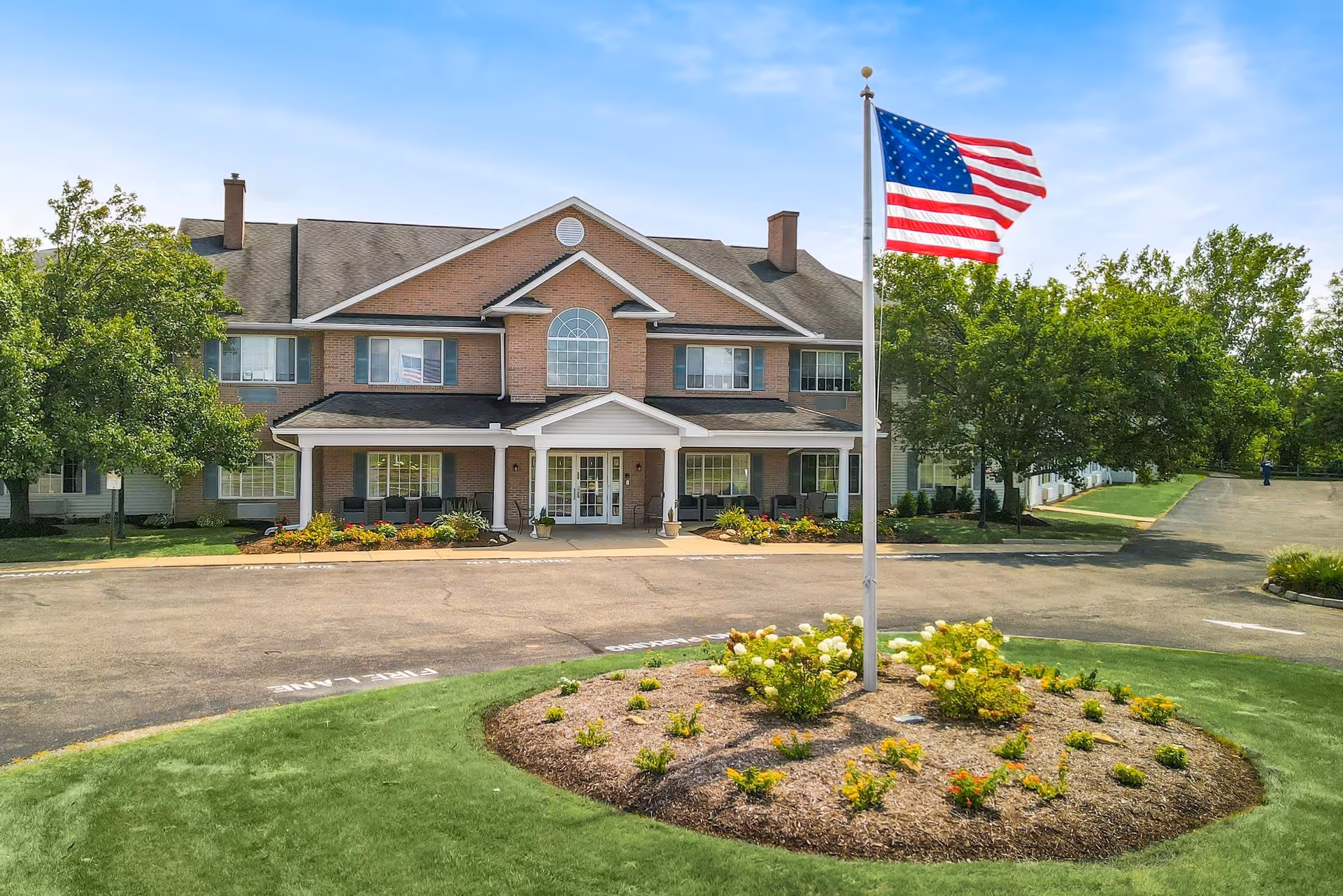 Front exterior view of Emeritus at Canton senior living facility with a circular driveway, landscaped flower bed with an American flag in the center, and trees surrounding the building under a blue sky.