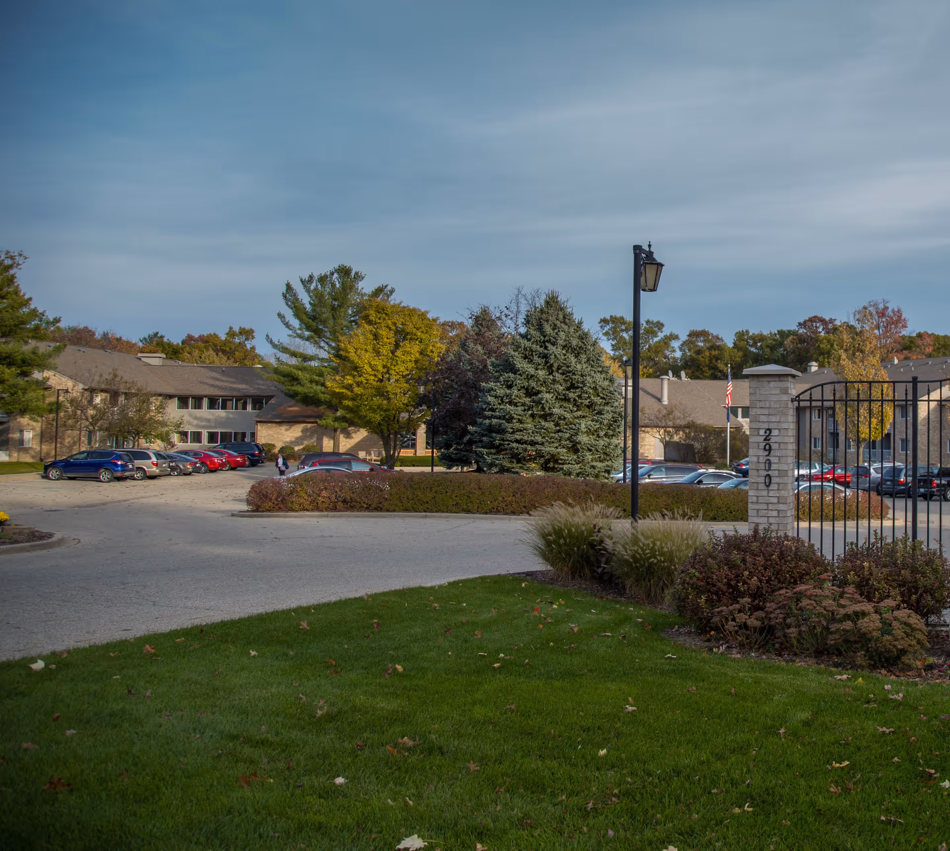 View of the entrance and parking area of a retirement community with a gated entrance, lamp post, green lawn, bushes, trees, and a two-story building in the background under a cloudy sky.