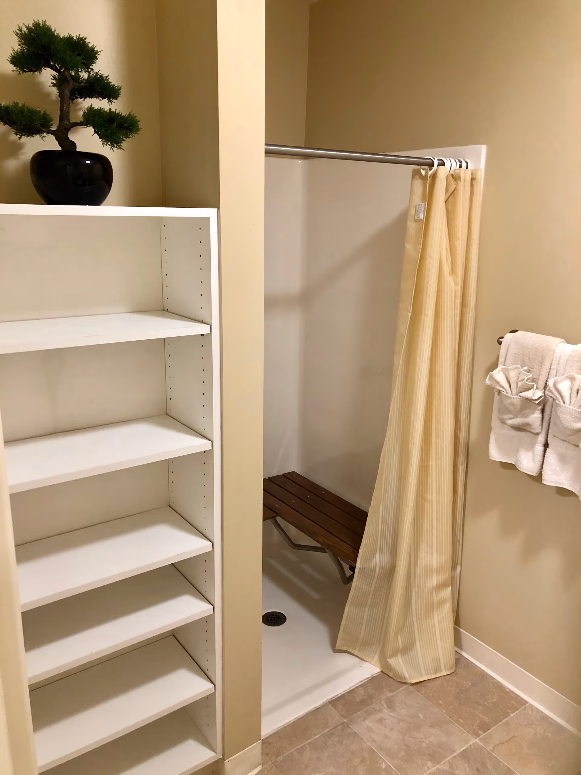 Accessible bathroom with a shower stall that has a wooden bench and striped curtain, a towel rack with folded towels, and adjacent white shelving topped with a small potted plant.