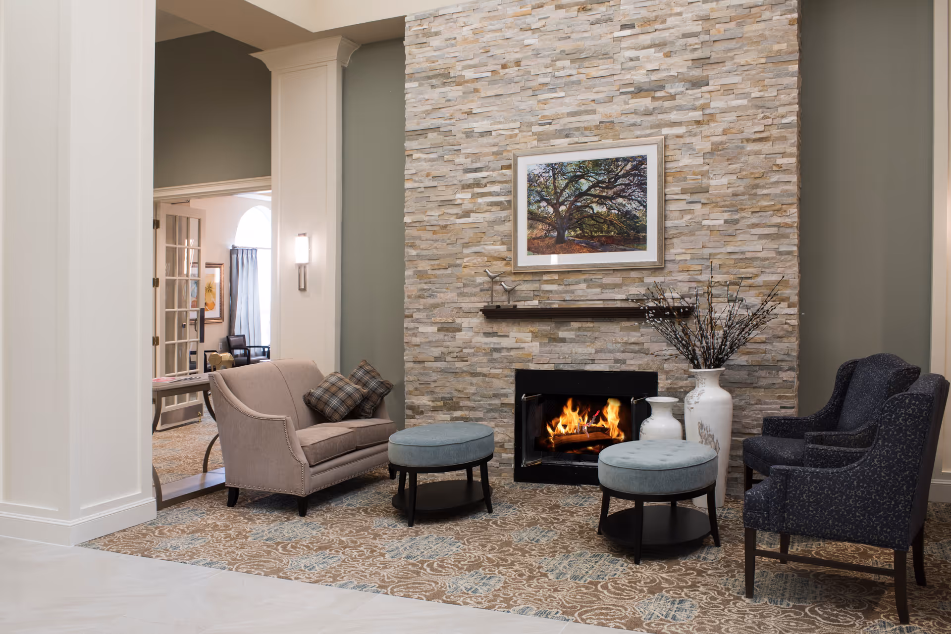A cozy living room area with a stone fireplace featuring a burning fire. Above the fireplace is a framed picture of a large tree. The room has a beige loveseat with two plaid pillows, two blue round ottomans, and two dark upholstered armchairs. A large white vase with decorative branches is placed next to the fireplace. The floor is covered with a patterned carpet, and there is a glimpse of another room through an open doorway.