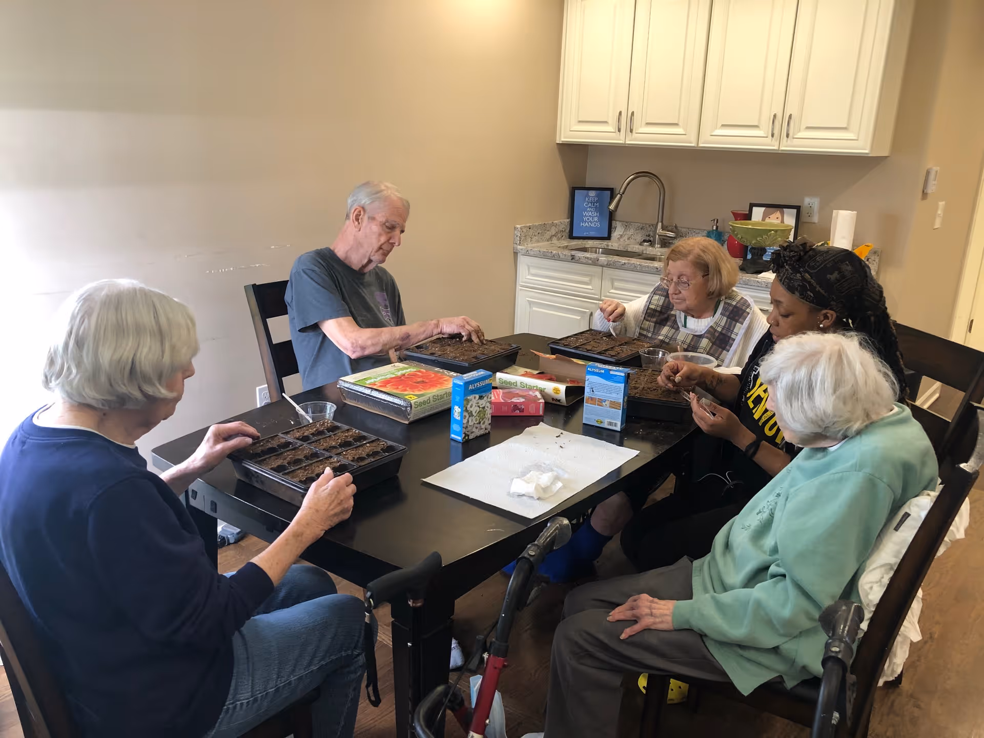 Five elderly individuals sitting around a table in a room with a kitchenette, engaging in a gardening activity by planting seeds in small trays filled with soil. The table has seed packets and gardening supplies on it.