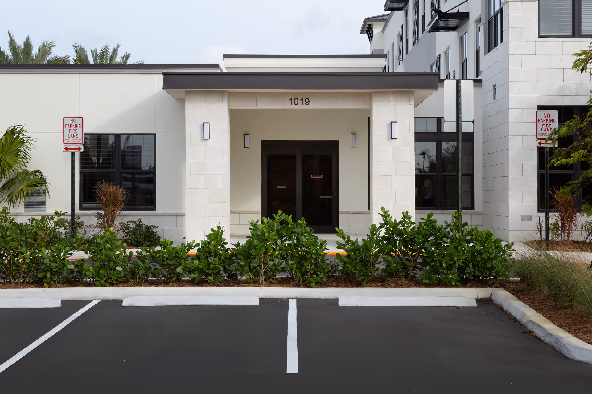 Front entrance of a modern building with the number 1019 above the doorway, flanked by two columns and wall-mounted lights. There are green shrubs in front of the entrance and two 'No Parking Fire Lane' signs on either side. The foreground shows an empty parking area with clearly marked spaces.