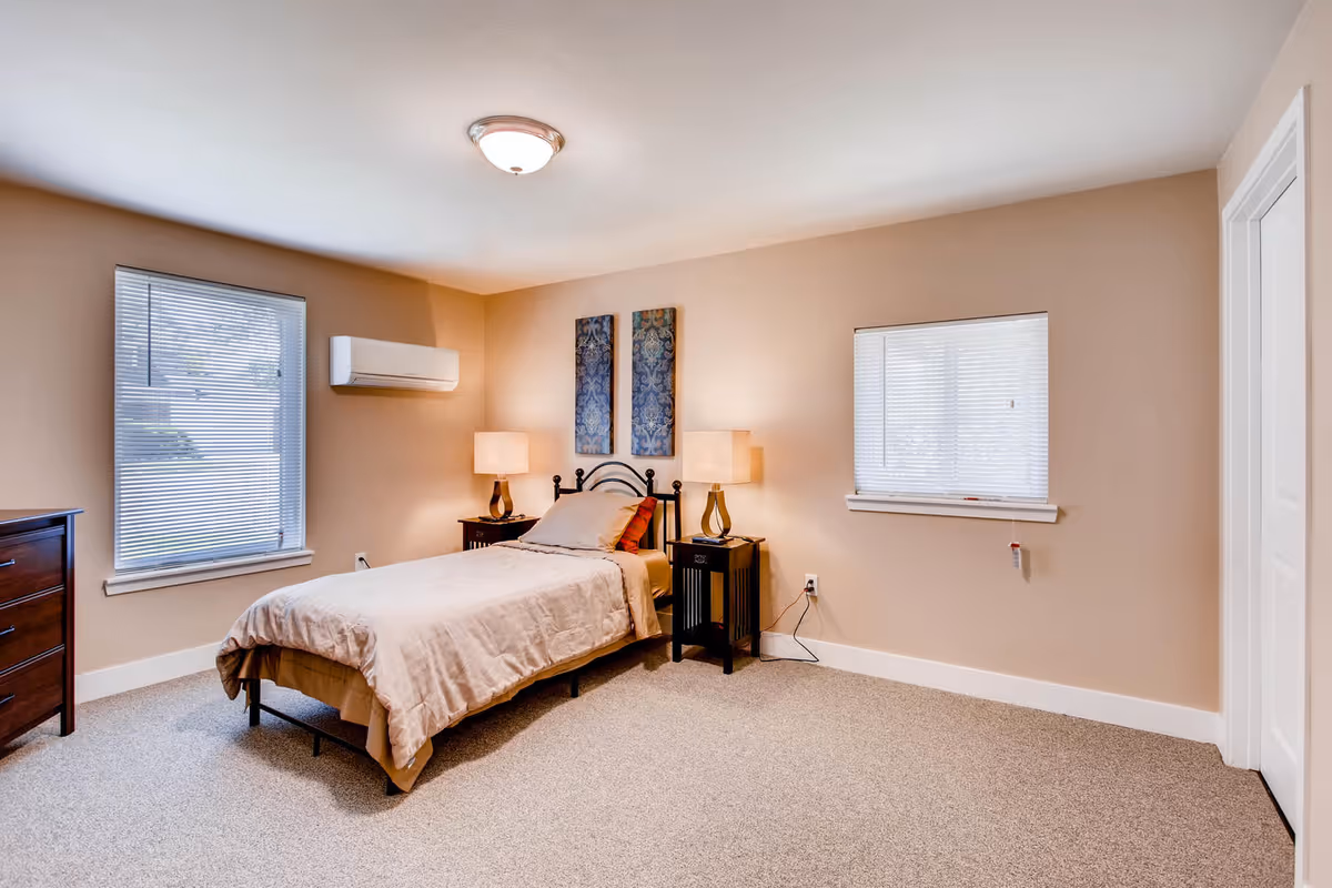 A neatly arranged bedroom with a single bed covered in beige bedding, two bedside tables with lamps, two decorative wall hangings above the bed, two windows with blinds, a wooden dresser, and a wall-mounted air conditioning unit.