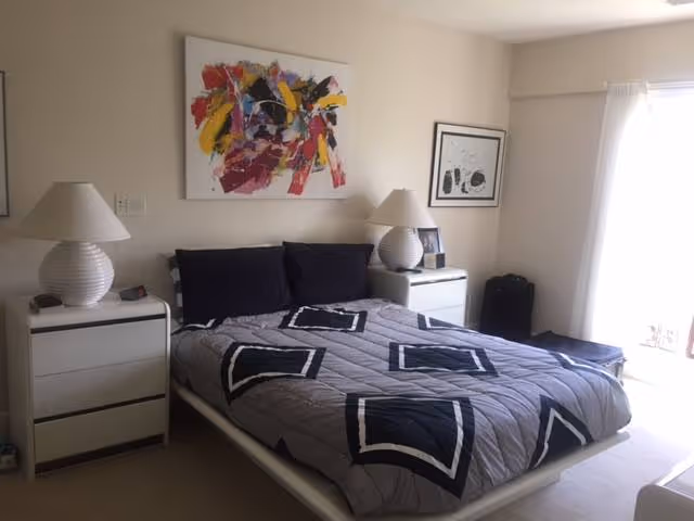 A bedroom with a bed covered in a gray quilt with black and white geometric patterns. There are two black pillows on the bed. On each side of the bed, there are white nightstands with white lamps. Above the bed hangs a colorful abstract painting, and another framed artwork is on the adjacent wall. A window with white curtains allows natural light into the room.