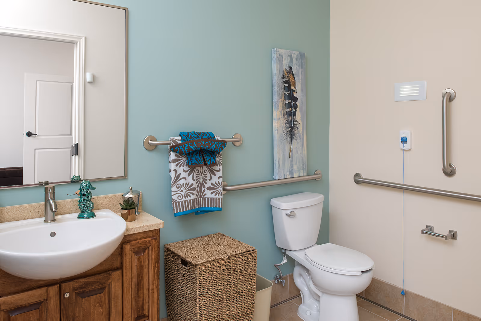 A clean and accessible bathroom featuring a white toilet with grab bars on the walls, a wicker laundry basket, a sink with a wooden cabinet, a large mirror, and decorative towels hanging on a towel bar. The walls are painted in light blue and beige tones, and there is a feather artwork hanging above the toilet.