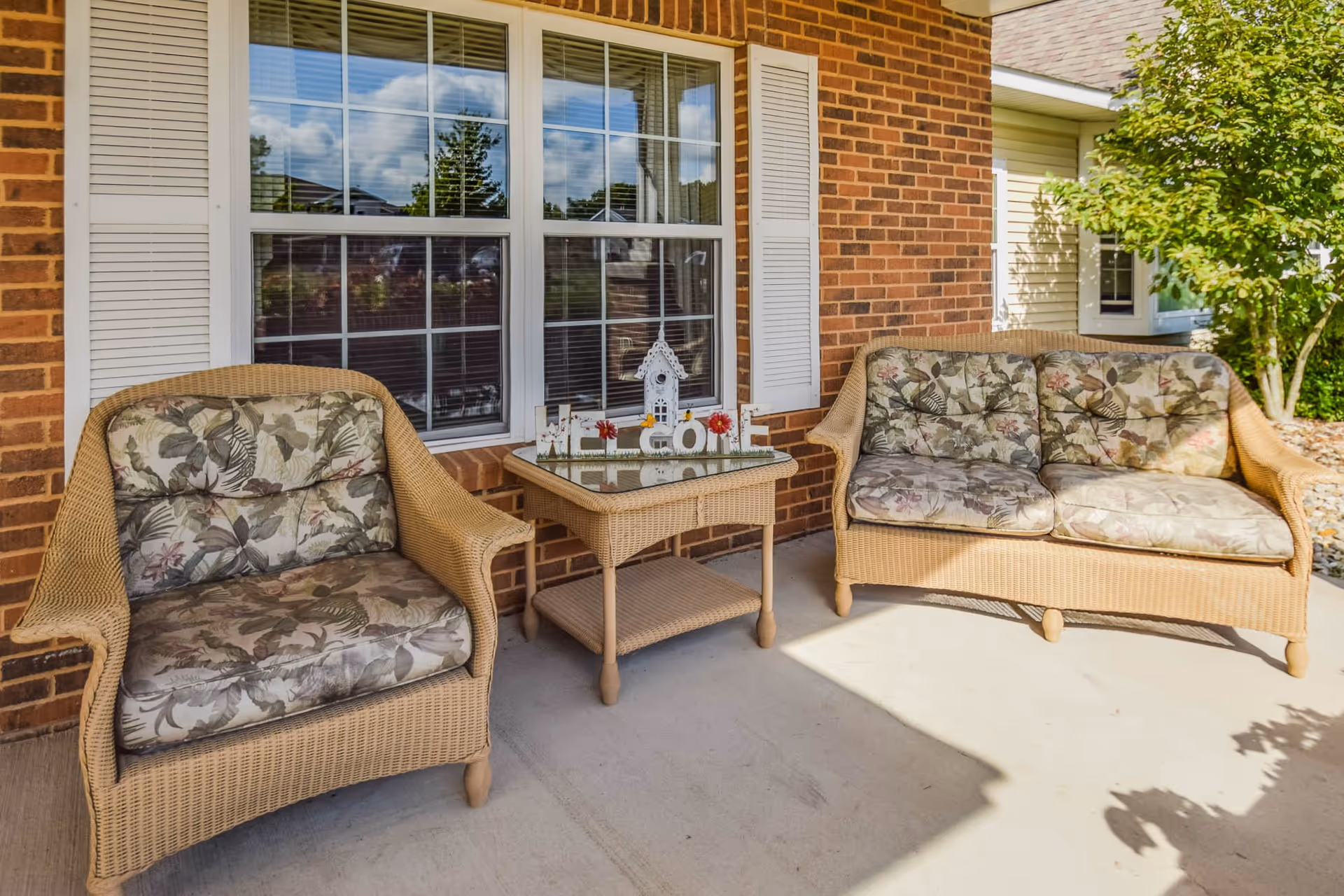 Outdoor patio area with two wicker chairs and a wicker loveseat, all with floral patterned cushions. A small wicker table with a glass top sits between the chairs, decorated with a small birdhouse and the word 'WELCOME'. The background shows a brick wall with a window and white shutters, and some greenery on the right side.