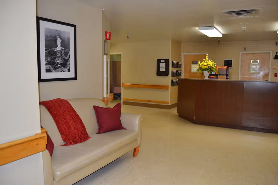 Interior view of a senior living facility reception area featuring a beige couch with red pillows and a red throw blanket. A framed black and white photo of the Statue of Liberty hangs on the wall above the couch. In the background, there is a wooden reception desk with a vase of yellow flowers and some office supplies. The floor is light-colored, and the walls are painted in neutral tones.