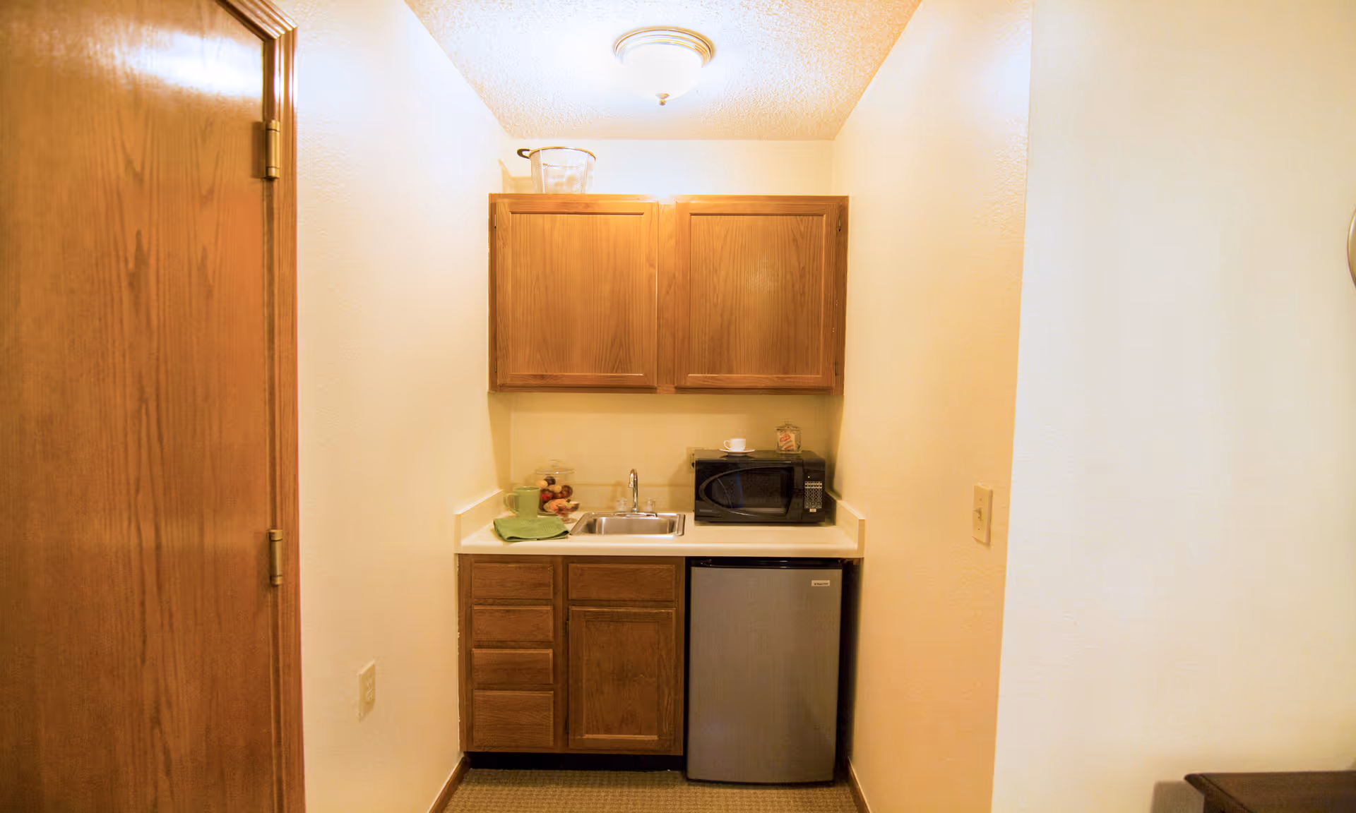 Small kitchenette with wooden cabinets, a sink, microwave, and mini fridge between beige walls.