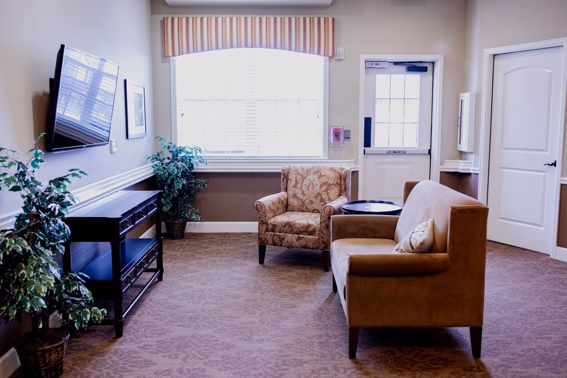 A bright communal seating area with a sofa and patterned armchair facing a wall-mounted TV, plants, and a windowed door.