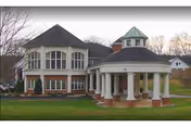 Exterior view of a two-story building with large windows and a covered entrance supported by white columns, surrounded by grass and trees with a few houses in the background.