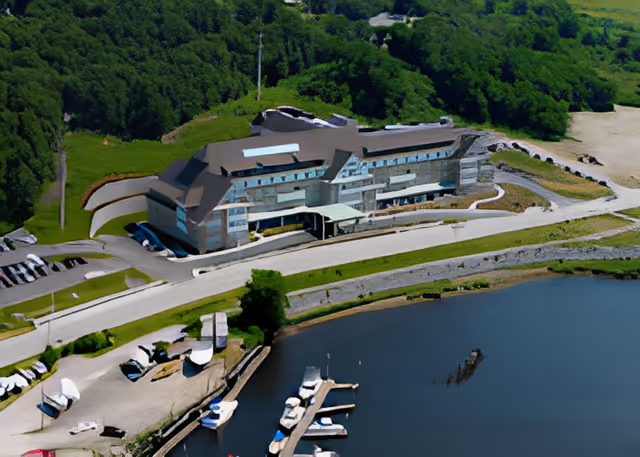 Aerial view of a large waterfront building surrounded by greenery and a parking area, with several boats docked in the adjacent water body.