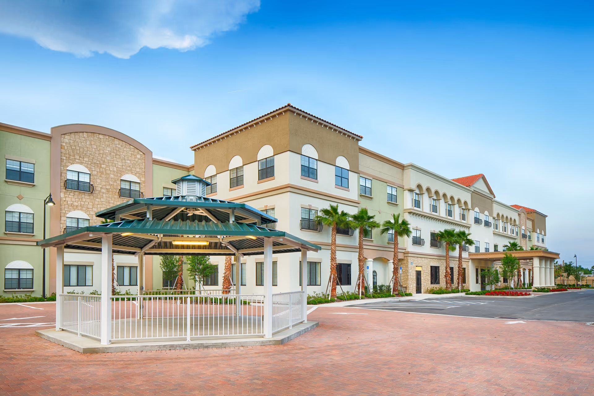 Exterior view of a multi-story senior living facility with a covered gazebo in the foreground, palm trees lining the building, and a clear blue sky above.