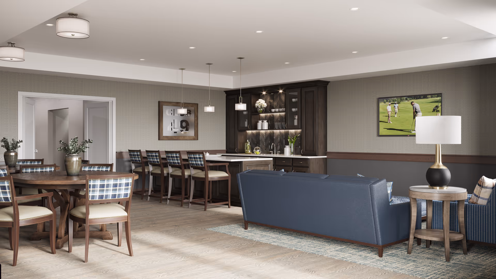 A spacious and well-lit common area in a senior living facility featuring a round wooden dining table with six chairs, a kitchen island with four bar stools, a dark blue sofa, an armchair, a side table with a lamp, and a wall-mounted TV showing a golf game. The room has light wood flooring, neutral-colored walls, and modern pendant lights above the kitchen island.