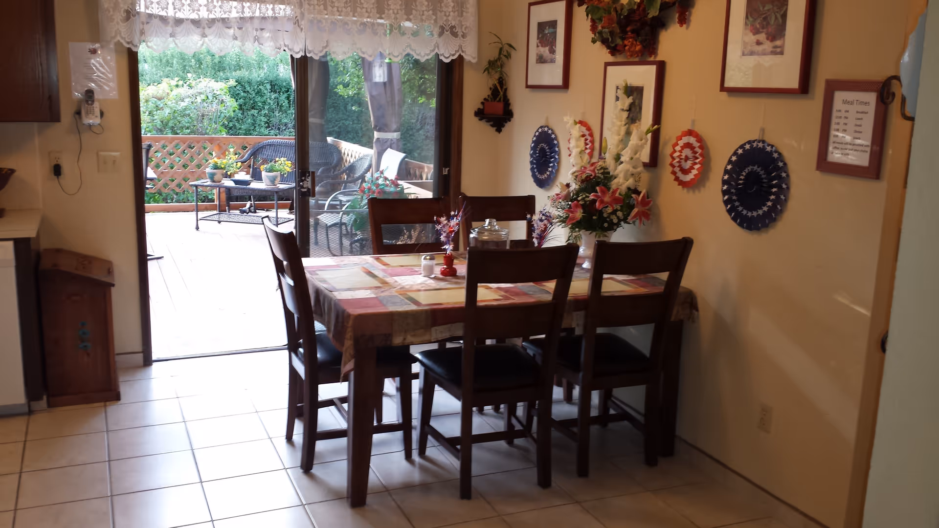 A dining area with a rectangular table covered with a patterned tablecloth and six wooden chairs. The table has decorative items including a flower vase and a jar. The room has tiled flooring and walls decorated with framed pictures and patriotic-themed wall hangings. A sliding glass door leads to an outdoor patio with wicker furniture and potted plants.