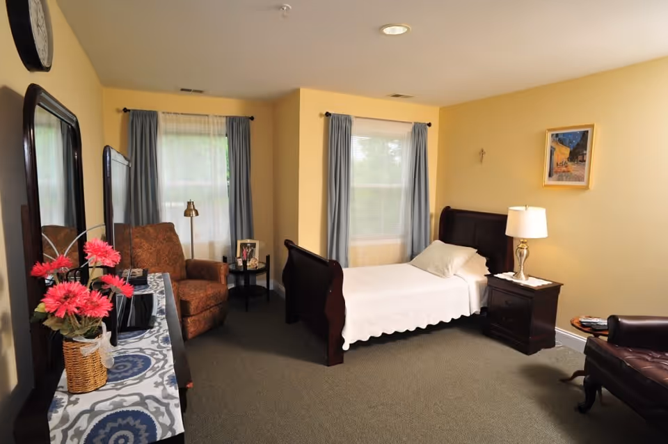 A cozy bedroom with a single bed covered in a white bedspread, a wooden nightstand with a lamp, two windows with blue curtains, a brown armchair, a dresser with a mirror, and a vase of pink flowers on top.