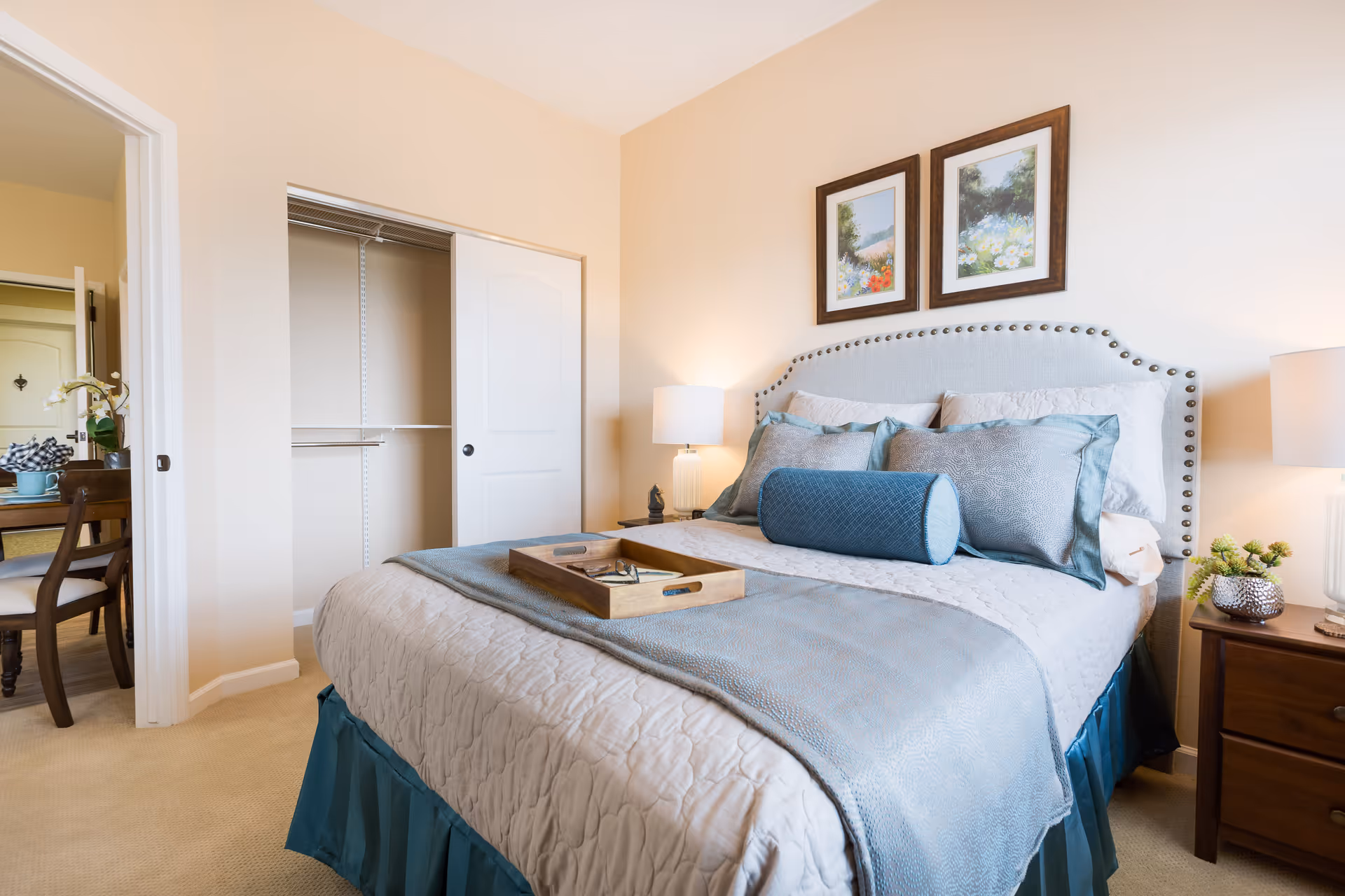 A neatly made bed with a light gray quilt, blue decorative pillows, and a blue bolster pillow in a bedroom. The bed has a light gray upholstered headboard with nailhead trim. On the bed is a wooden tray with a pair of glasses and a book. There are two framed floral paintings on the wall above the bed. A wooden nightstand with a lamp and a small plant is beside the bed. An open closet with sliding doors is visible, and a doorway leads to a dining area with a table and chairs.