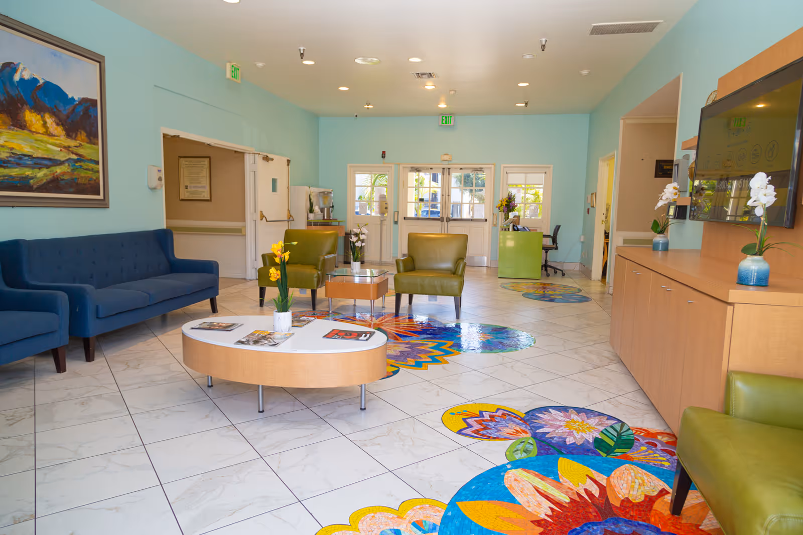 A bright and welcoming senior living facility lobby with light blue walls and white tiled floor featuring colorful floral mosaic designs. The room includes blue and green armchairs, a wooden coffee table with magazines and a small plant, a large wall-mounted TV, and a reception desk near the entrance doors. A landscape painting hangs on the left wall.